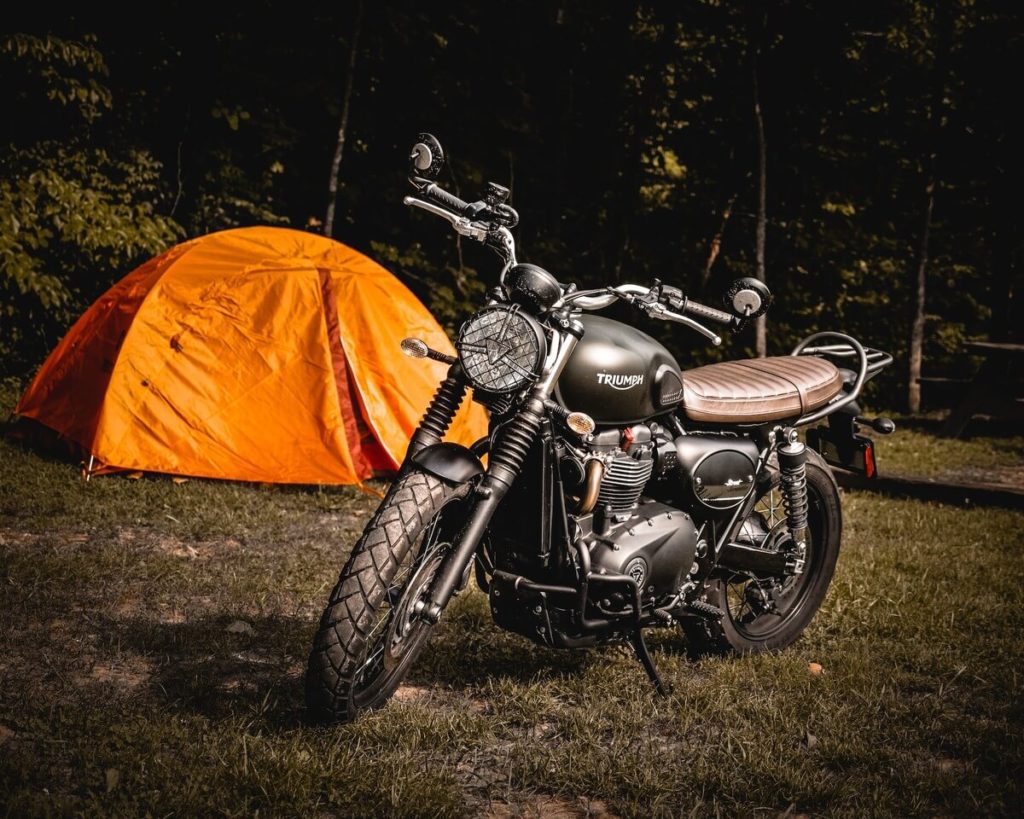 Image of someone motorcycle wild camping in Scotland with an orange tent in the background and a triumph motorcycle in the front.